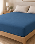 Lençol ajustável Stone azul a revestir o colchão numa cama com almofadas bege.