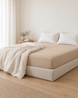 Lençol Ajustável Charming em Areia numa cama com sommier forrado, num quarto luminoso.